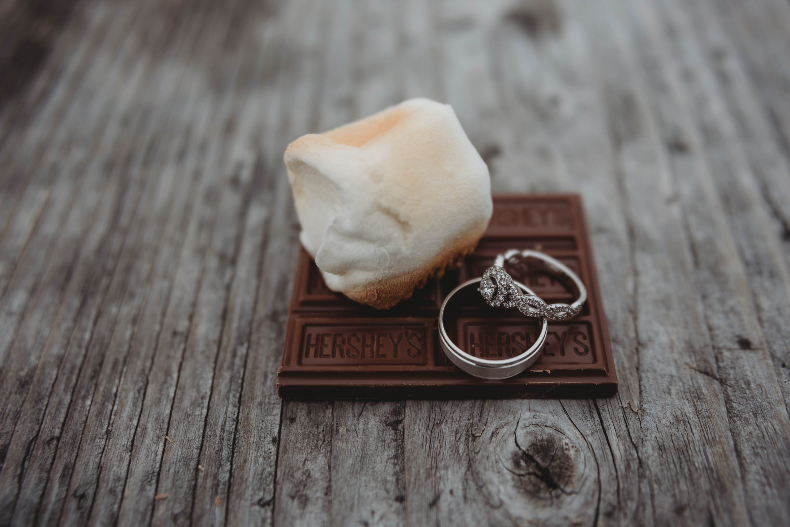 Wedding rings romantically sit next to a perfectly toasted marshmallow on a bar of chocolate.